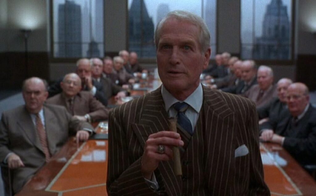 Man in business suit holding a cigar standing with his back to a long table with other businessmen seated around it.