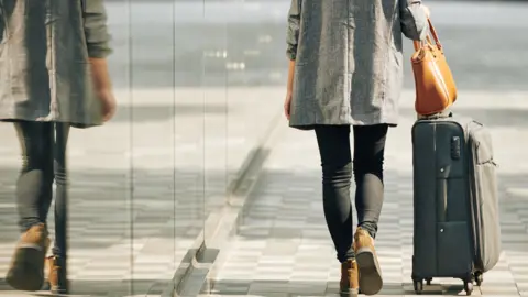 Getty Images A woman with her back to the camera wheeling a suitcase down the street.