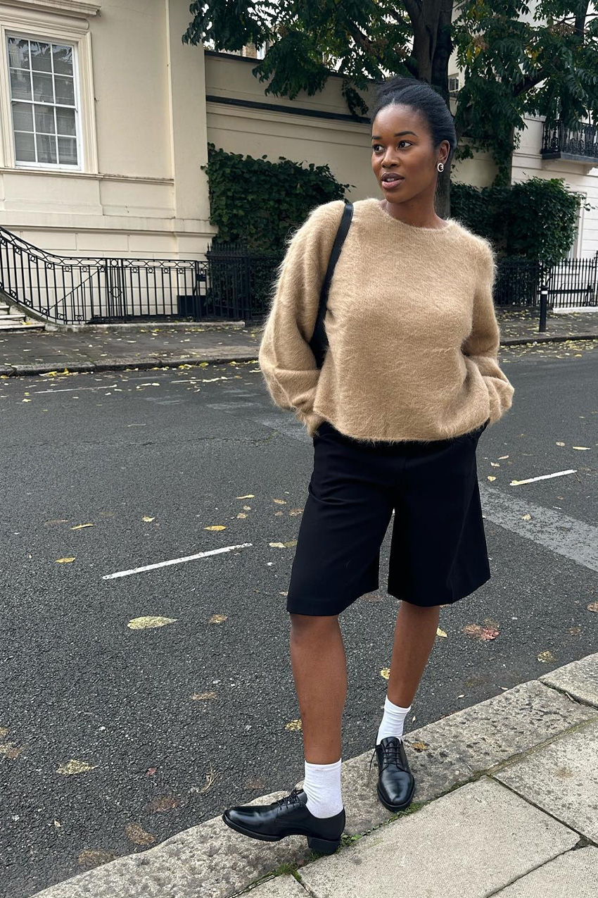 Shoe trends to wear with leggings, jeans, and skirts are shown in a street style photo of a woman standing on the sidewalk wearing silver earrings, a tan fuzzy crewneck sweater, a black shoulder bag, black bermuda shorts, white socks, and black oxford shoes